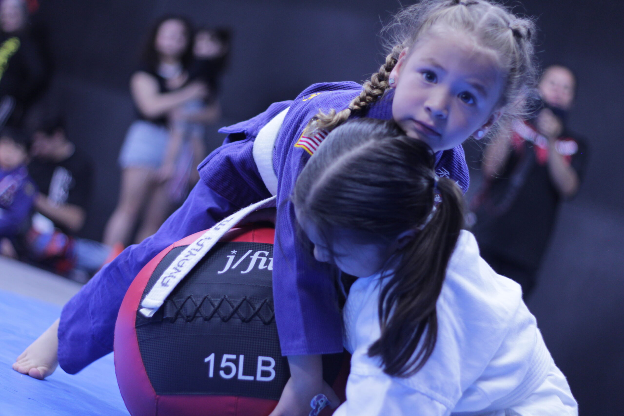 Two young girls in martial arts uniforms practice grappling on a blue mat